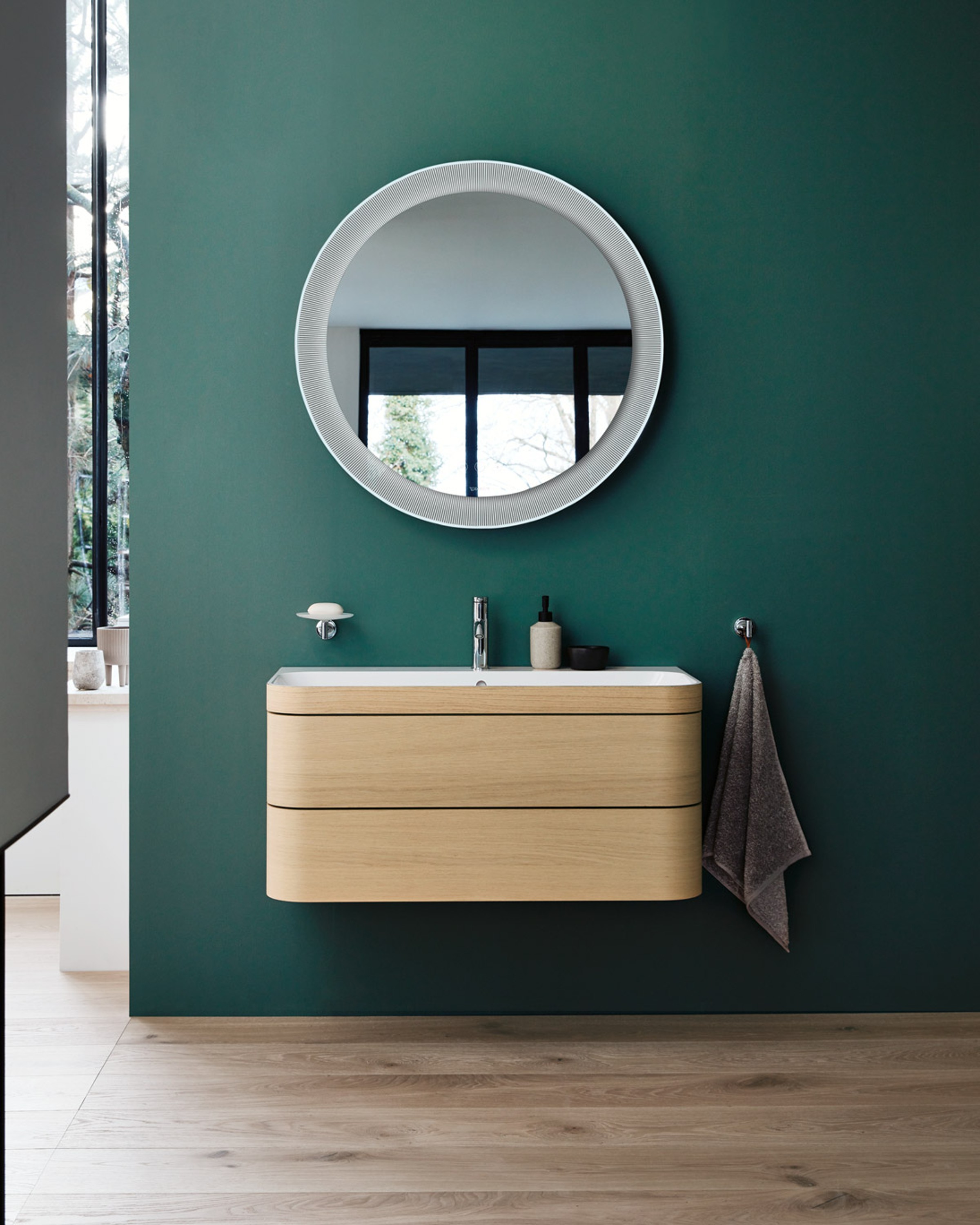 Duravit Happy D.2 vanity with round mirror hangs on green wall; towel rack next to it, trees visible outside.