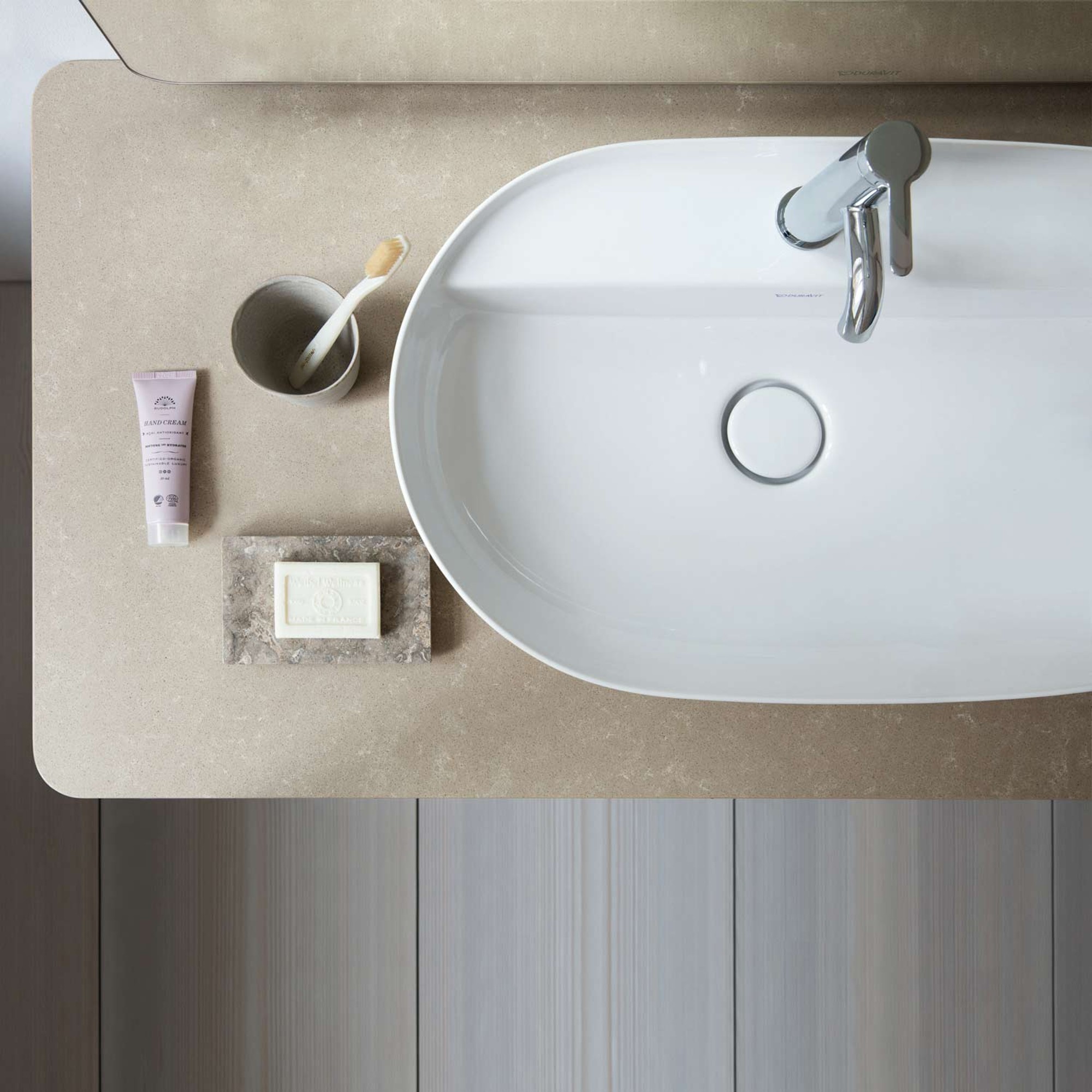 Duravit Balcoon Waschbecken hängt an brauner Wand, darunter Holzablage mit Handtüchern, daneben Vase. Dusch-WC im Hintergrund