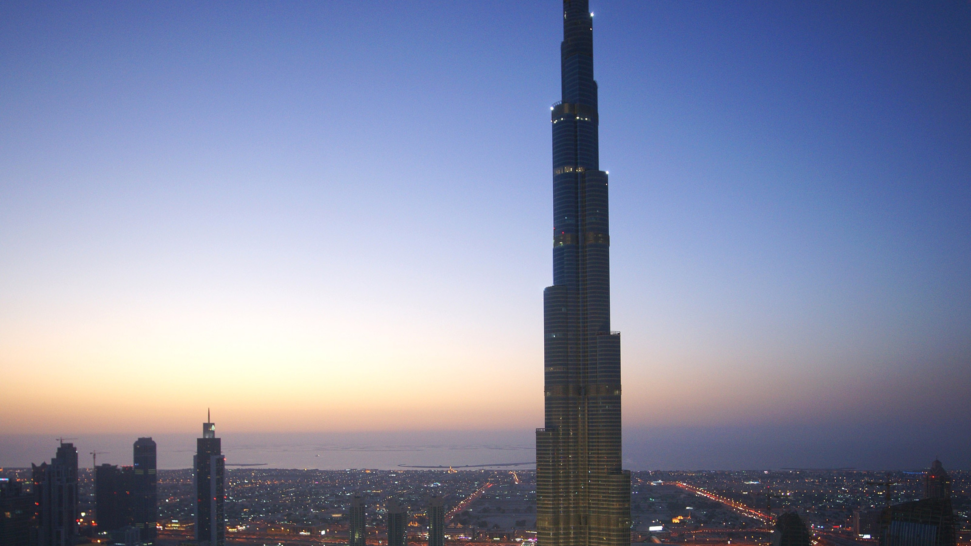 Burj Khalifa, Gebäudeansicht bei Sonnenuntergang mit beleuchteten Wasserspielen davor aus der Vogelperspektive