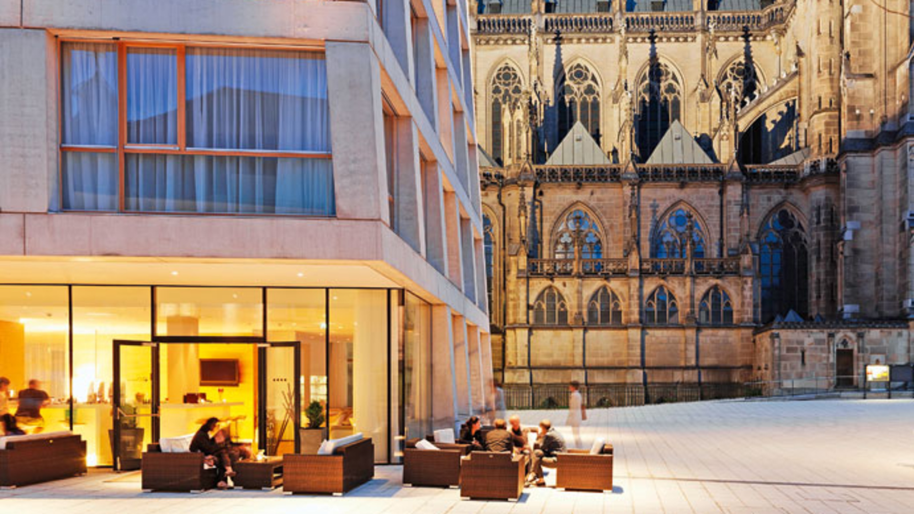 Hotel am Domplatz, exterior view of the building illuminated at dusk, with the hotel on the left and the cathedral on the right