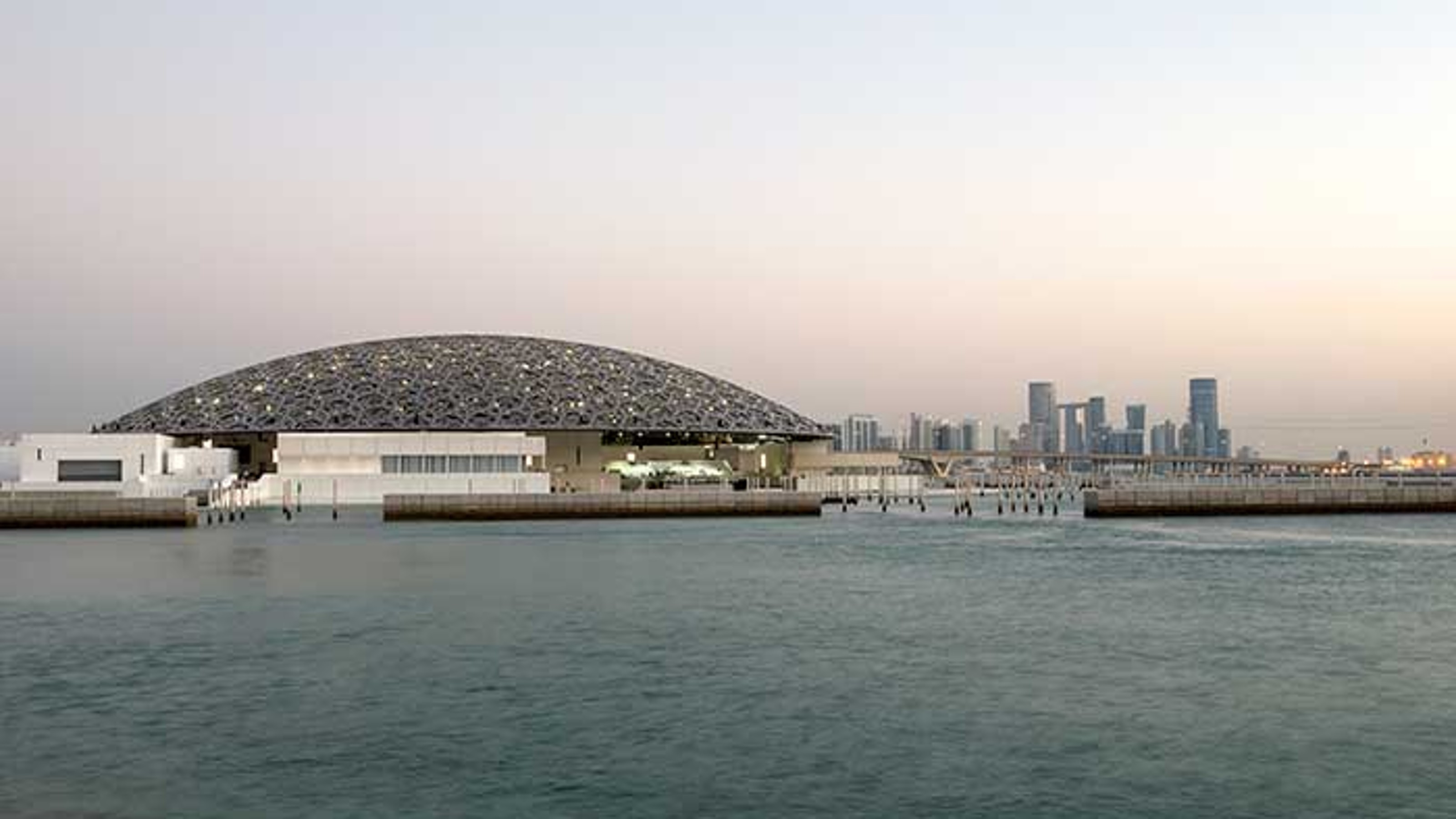 Louvre Abu Dhabi, panorama at dusk, building on the left, city on the right, water in the foreground