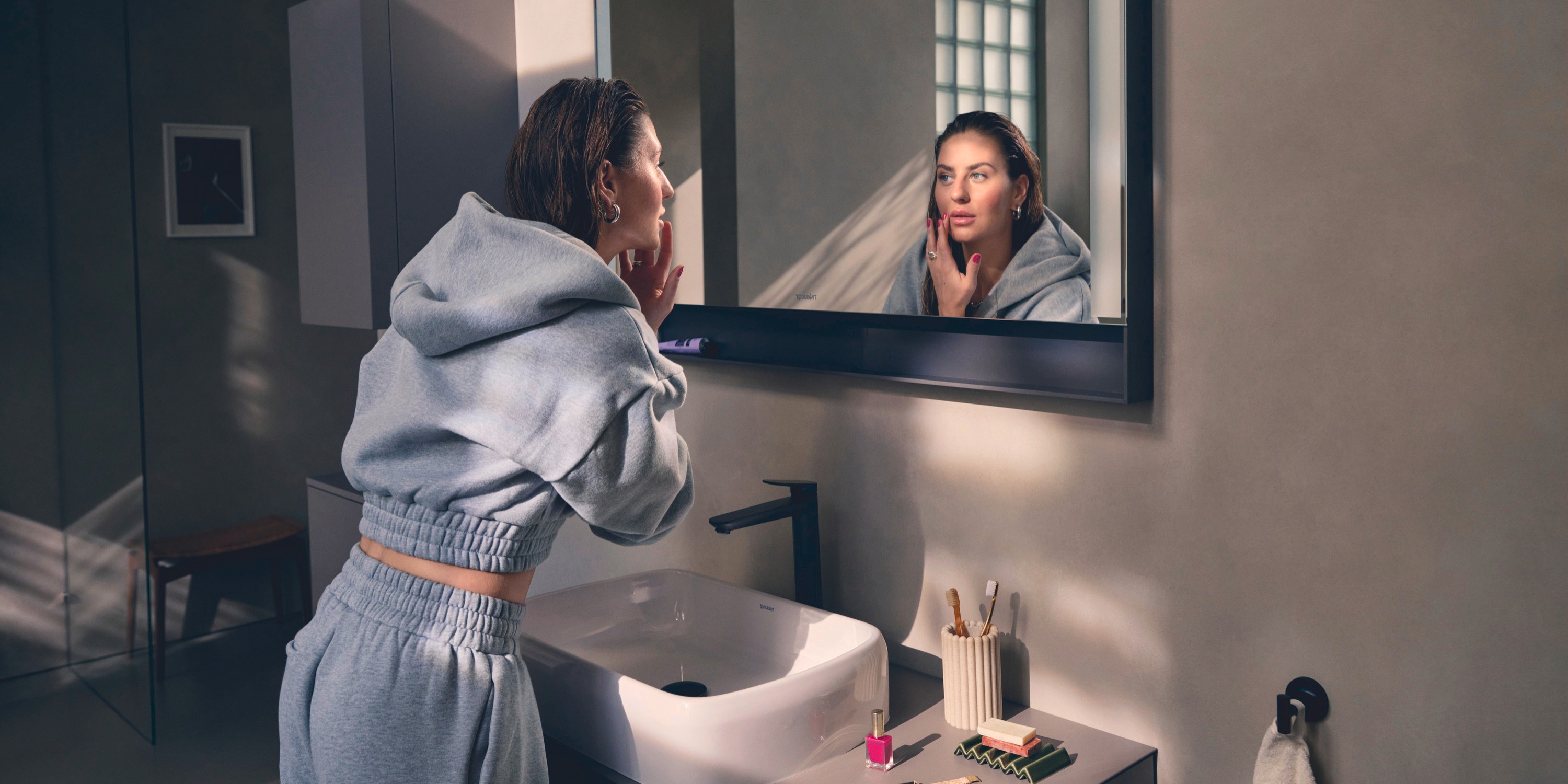 A woman is standing at a sink area from Duravit’s Studio F. A. Porsche Collection and applying makeup in the mirror