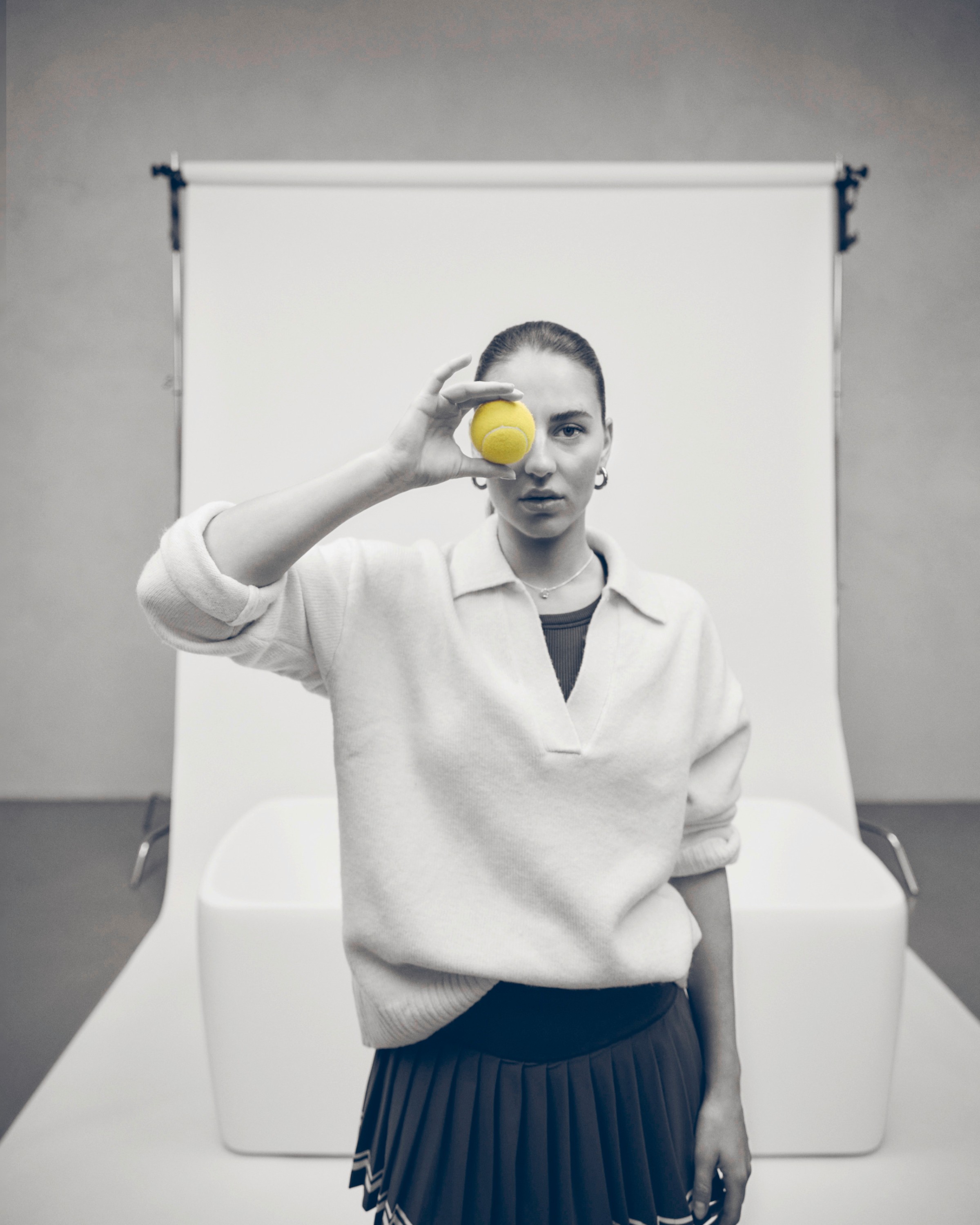 A woman is standing in a photo studio in front of a Bathtub, holding a tennis ball up to her eye 